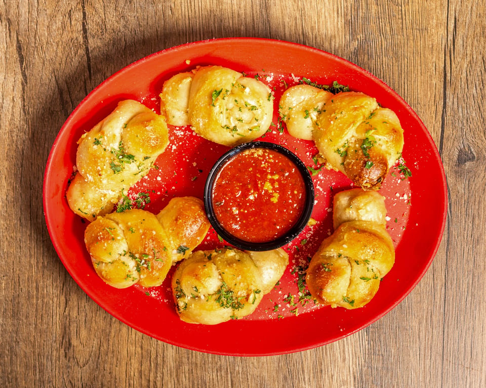 Golden garlic knots with parsley and parmesan cheese, surrounding a cup of marinara sauce on a red plate.