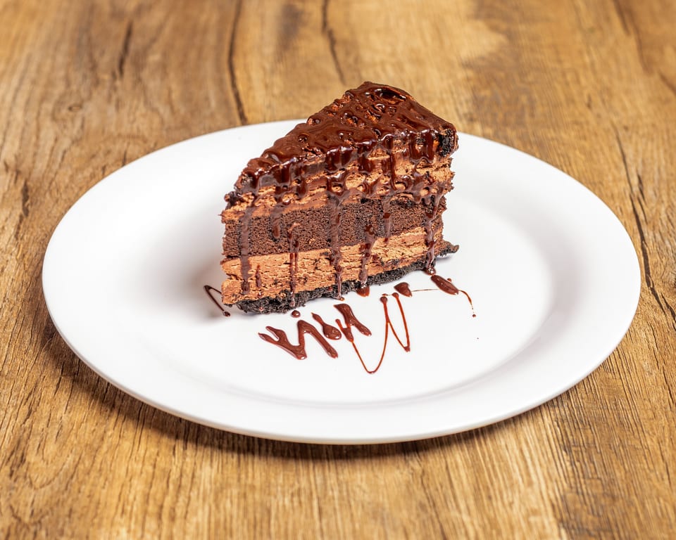 A slice of chocolate cake with multiple layers of frosting and chocolate drizzle on a white plate.