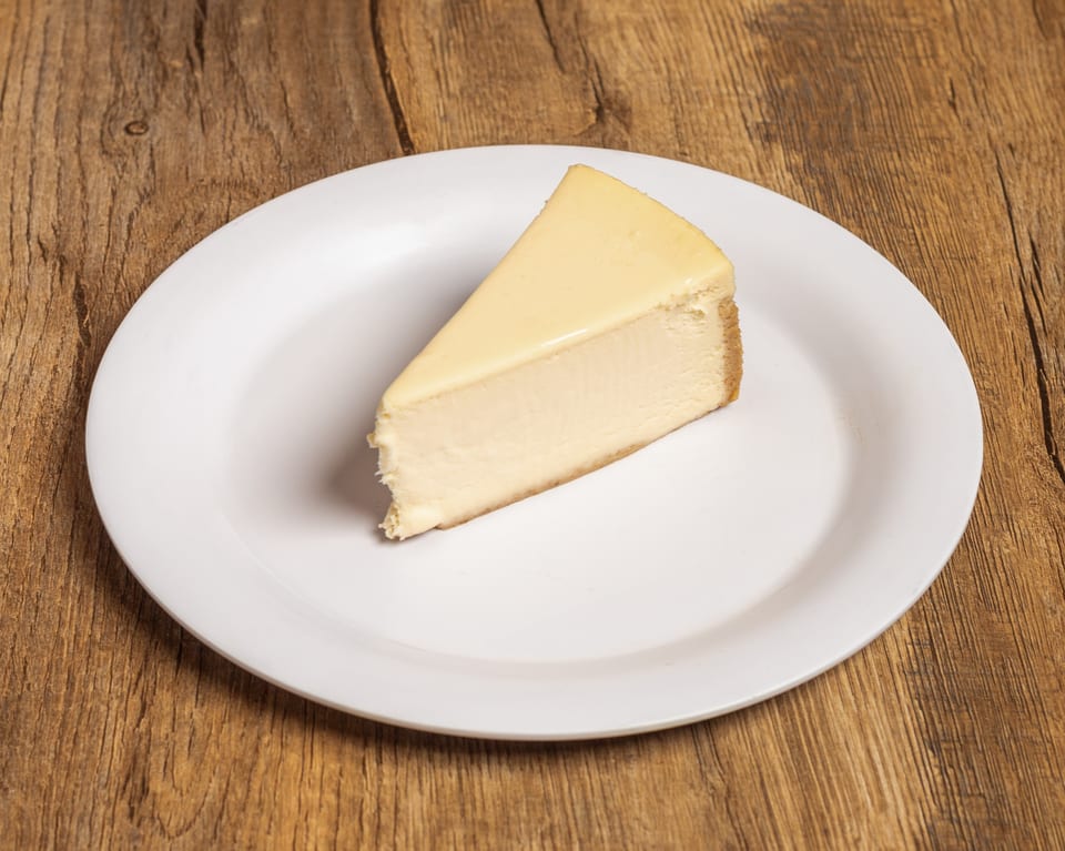 A single slice of plain cheesecake with a graham cracker crust on a white plate.