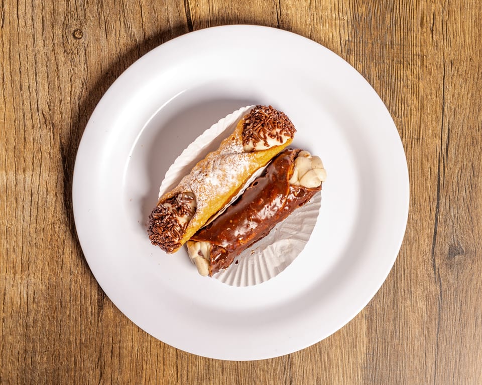 Two cannolis, one with chocolate sprinkles and one with chocolate glaze, on a white plate.