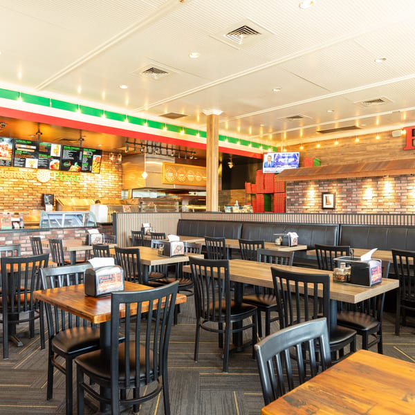 An empty, brightly lit dining room at Colonna's Pizza, featuring wooden tables, black chairs, and brick walls.