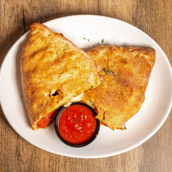 Two golden-brown calzones, one cut open to reveal melted cheese and pepperoni, with a side of marinara sauce.