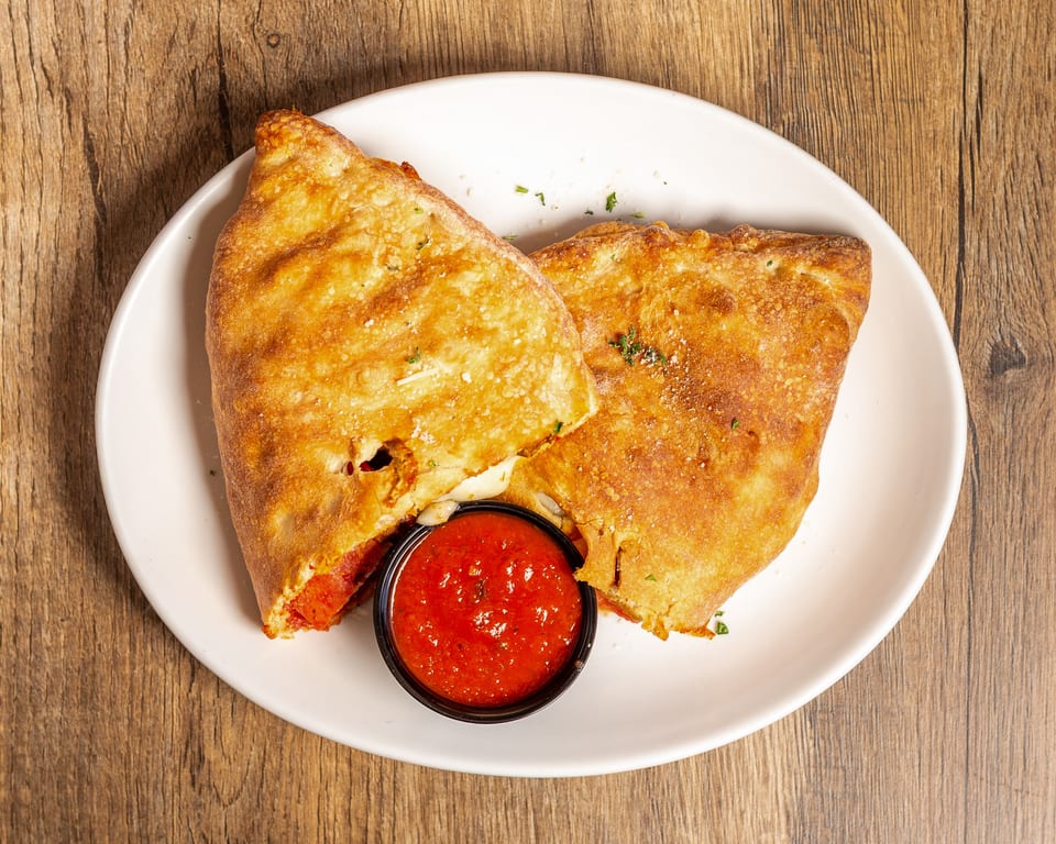 Two golden-brown calzones, one cut open to reveal melted cheese and pepperoni, with a side of marinara sauce.