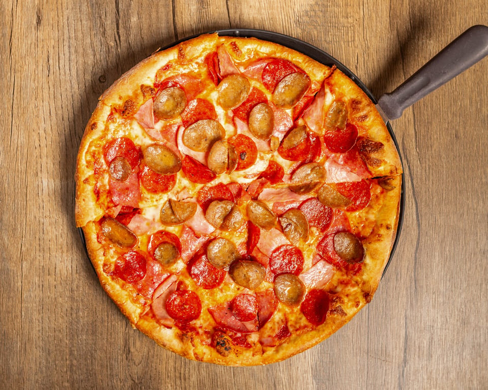 Overhead view of a whole pepperoni, ham, and sausage pizza on a dark pan.