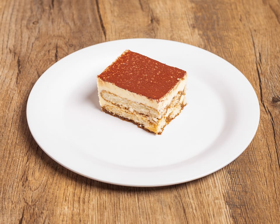 A rectangular slice of tiramisu with cocoa powder on top, served on a white plate.