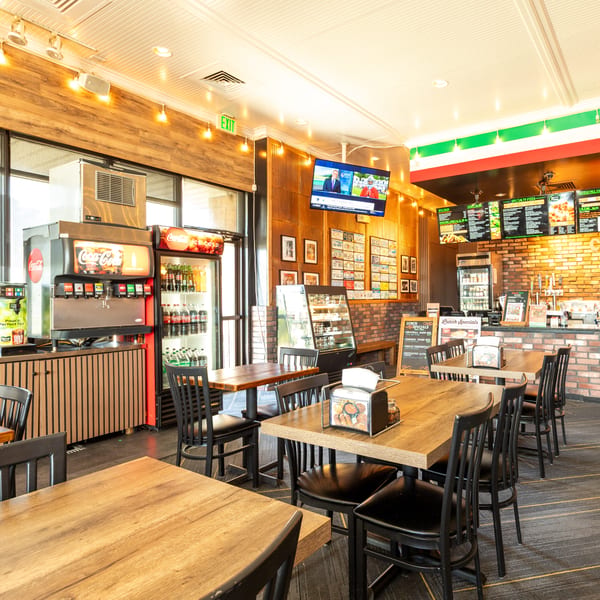 A brightly lit restaurant interior with wooden tables, black chairs, a soda fountain, and a menu board.