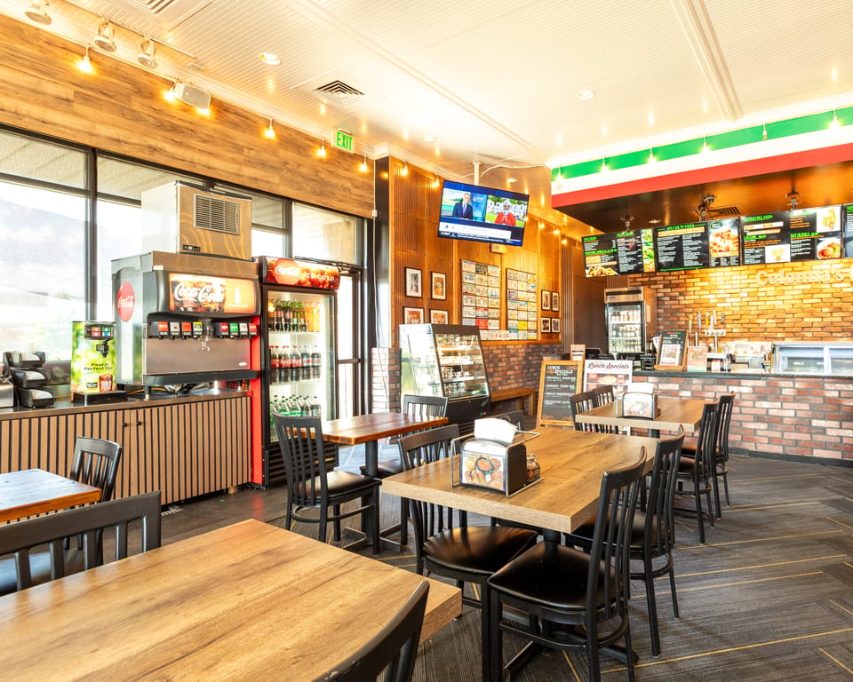 A brightly lit restaurant interior with wooden tables, black chairs, a soda fountain, and a menu board.