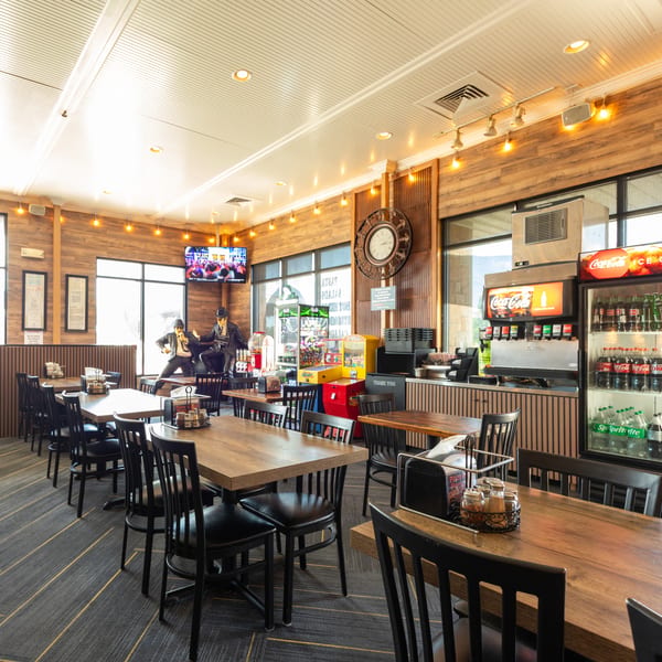 A brightly lit restaurant dining room with wooden tables, black chairs, and a soda fountain.