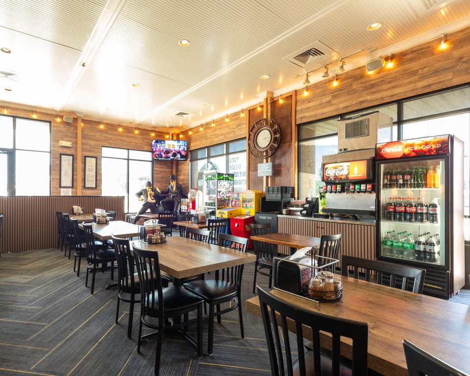 A brightly lit restaurant dining room with wooden tables, black chairs, and a soda fountain.